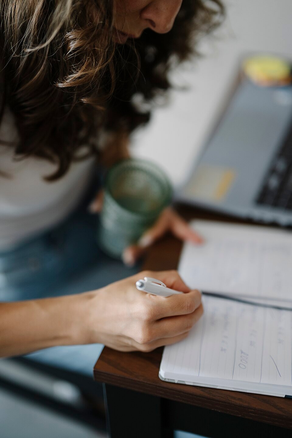 A person writing in a notebook while holding a drink near a laptop